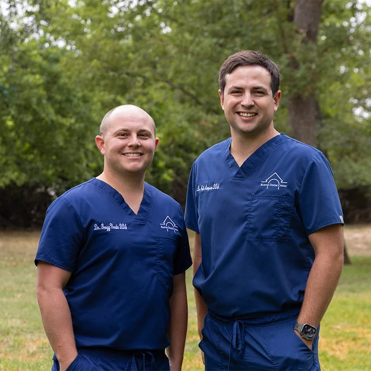 Dr. Gregg Vesta and Dr. Cole Simpson in navy scrubs, standing on the lawn under the oaks.