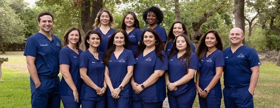 The whole Boerne Dental Center team in navy scrubs, standing in a line on the lawn under Hill Country oaks.